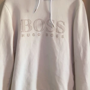 Vit hoodie från Hugo Boss - Säljer en stilren vit hoodie från Hugo Boss med logotypen tryckt på framsidan. Hoodien har långa ärmar och en klassisk huva med dragsko. 