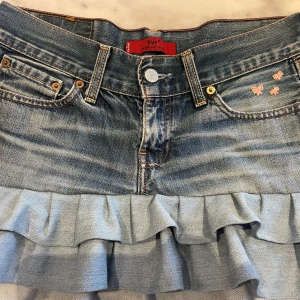 Lågmidjad jeanskjol med volang - Snygg jeanskjol med volangdetalj och gulliga rosetter på fickan. Skulle säga att den passar XS/S