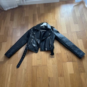 Svart skinnjacka från Pull&Bear - Snygg svart skinnjacka från Pull&Bear i storlek XS. Jackan har en klassisk bikerstil med dragkedjor och ett bälte i midjan. Perfekt för en cool och edgy look.