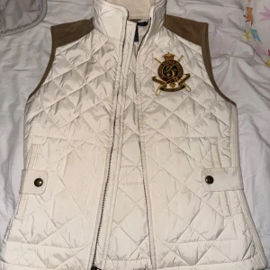 Vit quiltad väst från Ralph Lauren - Snygg vit quiltad väst från Ralph Lauren med bruna detaljer och ett broderat emblem på bröstet. Västen har dragkedja och knappar framtill samt en hög krage. Perfekt för en stilren look.