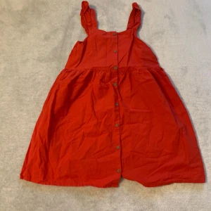 Röd klänning med knappar - Charmig röd klänning med knappar framtill och breda axelband. Perfekt för en somrig dag eller en avslappnad utflykt. Klänningen har en luftig passform och är knälång.