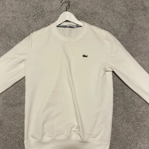 Vit tröja från Lacoste - Säljer en stilren vit tröja från Lacoste med det klassiska krokodilemblem på bröstet. Tröjan har långa ärmar och en rund halsringning. Perfekt för en avslappnad och snygg look.
