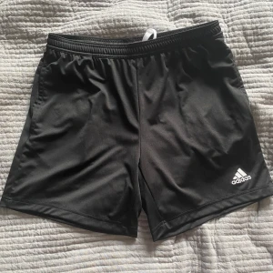 Svarta träningsshorts från Adidas - Säljer ett par svarta träningsshorts från Adidas. De har en elastisk midja med vit snörning för justerbar passform. Perfekta för sport och träning. Adidas-loggan är diskret placerad på benet.