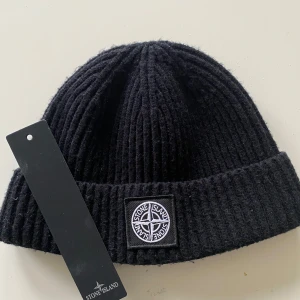 Svart mössa från Stone Island - Snygg svart ribbad mössa från Stone Island med uppvikt kant och logotyp framtill. Tillverkad i Italien av 100% bomull, perfekt för att hålla värmen med stil.