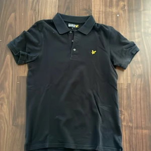 Svart pikétröja från Lyle & Scott - Säljer en stilren svart pikétröja från Lyle & Scott med klassisk krage och knappar framtill. Tröjan har en broderad gul logotyp på bröstet och korta ärmar. Perfekt för en avslappnad men ändå snygg look. Storlek L