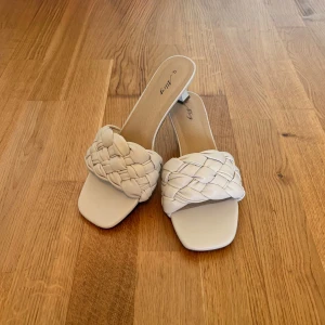 Vita flätade sandaler med låg klack - Snygga sandaler från Ally i vit färg med bred, flätad rem över foten och fyrkantig tå. De har en låg klack och öppen tå, vilket ger en trendig och fräsch look. Perfekta för dig som gillar stilrena och moderna skor med det lilla extra.