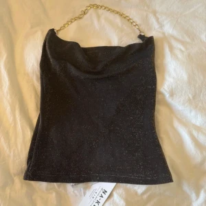 Svart glittrig tubtopp med guldkedja från NA-KD - Supersnygg svart tubtopp från NA-KD med glittrigt tyg och en chunky guldfärgad kedja som axelband. Toppen har en cool draperad halsringning och är croppad i modellen. Perfekt för att sticka ut med lite extra bling på festen!