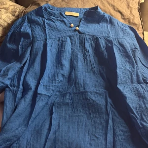 Blå långklänning från Freequent XS - Snygg blå långklänning från Freequent i storlek XS. Klänningen har en luftig och skrynklig bomullskänsla, v-ringad hals med två pärlemorknappar och långa, vida ärmar. Perfekt för dig som gillar en chill och avslappnad stil.