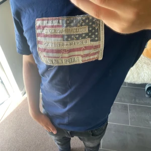 Blå t-shirt Ralph Lauren Denim & Supply - Snygg mörkblå t-shirt från Ralph Lauren Denim & Supply med ett stort patch på bröstet som har amerikanska flaggan och textdetaljer. Klassisk rund hals och korta ärmar. Tillverkad i mjuk bomull, perfekt för en chill och avslappnad stil.