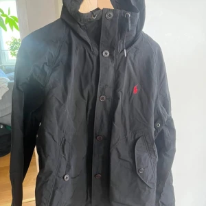Svart vindjacka från Polo Ralph Lauren - Svart vindjacka från Polo Ralph Lauren med röd broderad logga på bröstet. Jackan har huva med snörning, knappstängning framtill och praktiska fickor med knappar. Den har även en dragkedja bakom knapparna