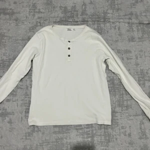 Vit långärmad tröja från Skill - Vit långärmad tröja från Skill med rund hals och tre svarta knappar framtill. Tröjan är gjord i mjuk bomull och har en clean, enkel look som funkar till många olika stilar. Perfekt för dig som gillar basic och bekvämt.