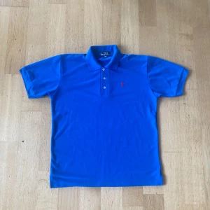 Blå polo t-shirt från Ralph Lauren - Snygg blå polo t-shirt från Ralph Lauren med klassisk krage och tre knappar framtill. T-shirten har korta ärmar och det ikoniska röda Polo-logot på bröstet. Tillverkad i mjuk bomull som är skön mot huden och passar perfekt till jeans eller shorts.