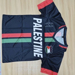 Palestina fotbollströja svart röd grön - Snygg fotbollströja med svart bas och trycket PALESTINE i vitt på framsidan. Röd och grön rand går vertikalt över tröjan, samt emblem med palestinska flaggan. Röd detalj på ärmar och krage. Materialet är glansigt polyester, perfekt för match eller träning.