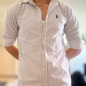 Randig skjorta från Polo Ralph Lauren - Snygg vit skjorta med mörkblå vertikala ränder från Polo Ralph Lauren. Klassisk krage, långärmad och med knappar hela vägen. Ikoniska Polo-loggan broderad på bröstet. Skjortan är gjord i mjuk bomull och har en normal passform.
