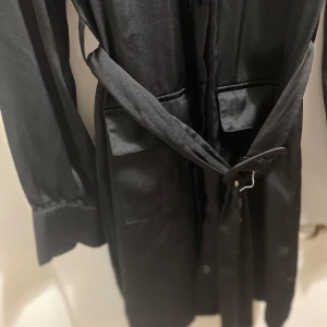 Svart skjortklänning med bälte Bik Bok - Snygg svart skjortklänning från Bik Bok i satinliknande polyester. Klänningen har långa ärmar med manschett, fyra stora fickor framtill, knappar hela vägen och ett matchande bälte i midjan. Perfekt för dig som gillar utility-vibe och stilrena detaljer. Storlek Xs men är mer som en S. 