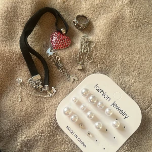 Mix av smycken med pärlor och hjärta - Säljer ett smyckespaket med silverfärgad ring, halsband med rött glittrande hjärta på svart band, flera tunna kedjor och ett set med pärlörhängen i olika storlekar. Perfekt för dig som gillar att mixa och matcha accessoarer.