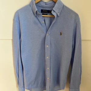 Blå skjorta från Polo Ralph Lauren - Snygg ljusblå skjorta från Polo Ralph Lauren med klassisk button-down krage och broderad logga på bröstet. Skjortan har långa ärmar, vita knappar och är tillverkad i mjuk bomull. Perfekt för dig som gillar stilrena och tidlösa plagg.