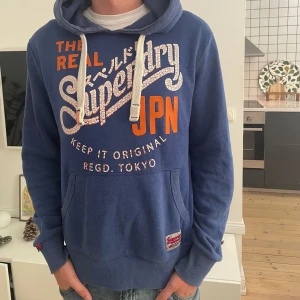 Blå Superdry hoodie med tryck - Snygg blå hoodie från Superdry i storlek medium med stort vitt och orange tryck på bröstet och coola japanska detaljer. Tröjan har huva med vita snören, känguruficka och ribbade muddar. Perfekt för dig som gillar streetwear och vill sticka ut.