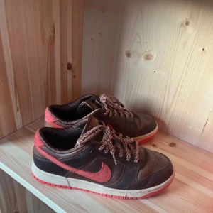 Nike Dunk Low i brunt och rött - Nike Dunk Low sneakers med brun ovandel i skinn och mocka, röda detaljer och Swoosh i textil. Skorna har quiltad insida, rund tå och platt sula. Snygga snören i brunt och vitt ger extra stilpoäng. Perfekt för dig som gillar unika färgkombinationer och streetwear.