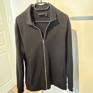 Svart zip-kofta  - Svart kofta från Jack & Jones med hel dragkedja framtill och hög krage. Tröjan har långa ärmar och två fickor på sidorna. Perfekt för dig som gillar enkel och stilren design. Materialet är mjuk bomull som känns skönt mot huden.