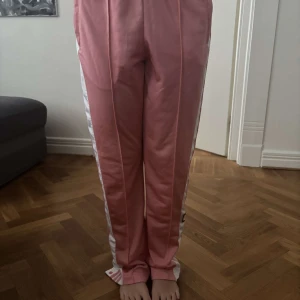 Rosa Adidas mjukisbyxor med knappar - Säljer ett par rosa Adidas mjukisbyxor med vita knappar längs sidan. Byxorna har tryckknappar hela vägen ner. Aldrig använda så dom är som nya. Är storlek 36 men upplevs mer som en 38.