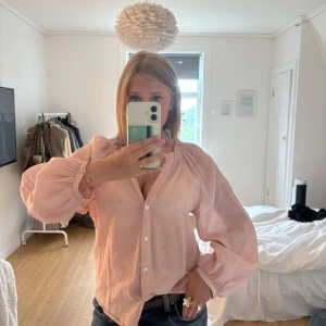 Ljusrosa blus med puffärm och knappar - Säljer en ljusrosa blus med v-ringning och vita knappar framtill. Blusen har långa puffärmar med resår vid ärmslut och är tillverkad i ett lätt, luftigt material som ger en skön känsla. 