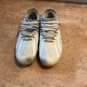Vita crazy fast fotbollsskor från Adidas - Säljer ett par vita fotbollsskor från Adidas med klassiska tre ränder på sidorna. Skorna har snörning och en låg profil med rund tå. Ovandelen är i syntetmaterial och sulan har dobbar för bra grepp på planen. Perfekt för dig som vill ha stil på träningen.