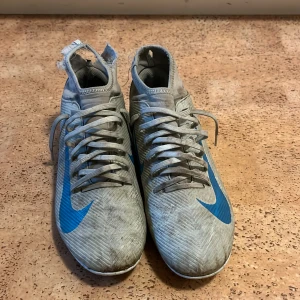 Nike Mercurial grå-blå fotbollsskor - Nike Mercurial fotbollsskor i grått med blå detaljer och stor Nike-logga på sidan och hälen. Skorna har snörning, platt sula med dobbar och en strumpliknande högre krage. Tillverkade i syntetmaterial med mönstrad yta för extra grepp på planen.