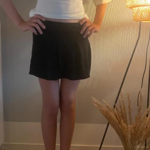 Svart kortkjol med veck - Snygg svart kortkjol med diskreta veck framtill som ger en cool och stilren look. Kjolen har en enkel design och sitter bekvämt i midjan. Perfekt för dig som gillar minimalistisk stil och vill ha en kjol som är lätt att matcha med olika toppar.