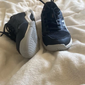 Svarta Asics inneskor strl 40.5 - Säljer ett par svarta Asics sneakers med vita detaljer och logga på sidan. Skorna har snörning, rund tå och platt sula. Ovandelen är i syntet och textil, vilket gör dem lätta och bekväma. Perfekta för dig som gillar sportig stil och vill ha ett par snygga skor till träning. Nypris:650
