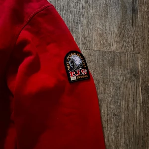 Röd sweatshirt från Parajumper - Säljer en röd sweatshirt från Parajumpers i storlek M. Tröjan har rund hals, ribbade muddar och ett snyggt PJS-märke på ärmen. Tillverkad i 100% bomull, perfekt för dig som gillar stilrena och bekväma plagg med lite attityd.