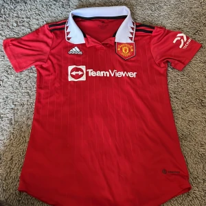 Manchester United röd matchtröja XS - Säljer en officiell Manchester United fotbollströja från Adidas i röd färg med vita och svarta detaljer. Tröjan har korta ärmar, vit krage med röd grafisk design och tunna vertikala ränder. Materialet är lätt och andas, perfekt för träning eller match. Skick 9/10