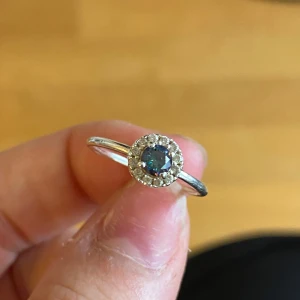 Silverring med blå sten och kristaller - Snygg och tunn silverring med en rund blå sten i mitten, omgiven av små glittrande kristaller. Ringen är stämplad med 925 och har en klassisk, elegant design som verkligen poppar på fingret. Perfekt för dig som gillar stilrena smycken med lite extra bling.