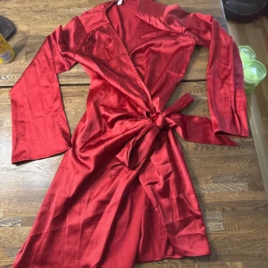 Röd omlottklänning med knytband - Snygg röd omlottklänning med långa ärmar och breda slag. Klänningen har ett knytband i midjan som ger en cool siluett och är gjord i ett glansigt satinliknande material. Perfekt för dig som vill sticka ut med en färgstark look.