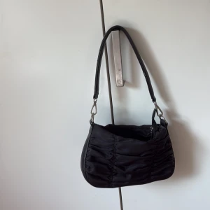 Svart axelväska med dragkedja - Snygg svart axelväska med rynkade detaljer och bred axelrem. Väskan har silverfärgade metalldetaljer och stängs med dragkedja. Materialet är en mix av syntet och skinn, vilket ger en cool och modern look. Perfekt storlek för det viktigaste.