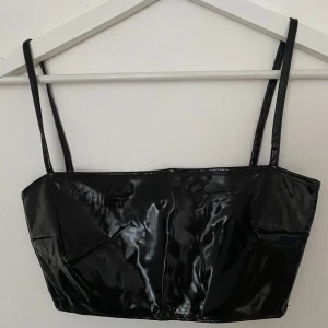 Svart glansig linne från NLY One - Svart topp från NLY One i storlek XXS. Toppens glansiga yta ger en edgy vibe och de smala axelbanden gör den extra snygg. Materialet är syntet och modellen är croppad, perfekt för att sticka ut med en cool look.
