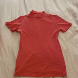 Röd polotopp från Zara - Snygg röd polotopp från Zara i ribbad bomull. T-shirten har korta ärmar och en hög krage. Tråden har gått av lite längst ner på baksidan men det är inget man lägger märke till💞. 