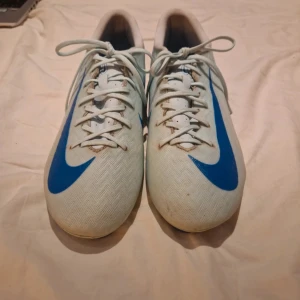 Nike Mercurial ljusblå och blå fotbollsskor - Nike Mercurial fotbollsskor i ljusblå och blå detaljer och stora Swoosh-loggor. Skorna har snörning och platt sula med fasta dobbar för gräsplan. Ovandelen är tillverkad i syntetmaterial med mönstrad struktur för bättre bollkontroll. Snygg och sportig design som sticker ut på planen.