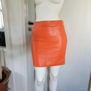 Orange skinnkjol från Boohoo - Säljer en snygg orange kort kjol från Boohoo i skinnimitation. Kjolen har en rak och tight passform med slät yta och enkel midja. Perfekt för dig som vill sticka ut med färg och gillar trendiga plagg. Storlek EU 38.