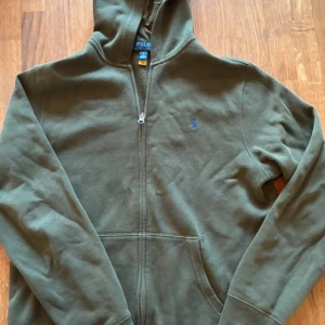 grön hoodie från Polo Ralph Lauren - Snygg grön hoodie från Polo Ralph Lauren i storlek XL i barnstorlek. Tröjan har huva, dragkedja framtill och en klassisk känguruficka. Liten blå logga broderad på bröstet. Perfekt för chill dagar och streetstyle.