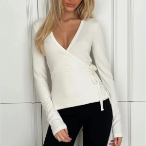 Vit omlottopp från Nelly - Vit omlottopp med lång ärm, v-ringad hals och ett stilrent knyt i midjan. Toppen är från Nelly i storlek M. Endast använd en gång. 