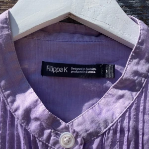 Ljuslila skjorta från Filippa K - Säljer en ljuslila skjorta från Filippa K med ståkrage och snygga veckade detaljer framtill. Skjortan har vita knappar hela vägen och är tillverkad i mjuk bomull. Långärmad och loose fit, perfekt för dig som gillar stilrena plagg med en twist.