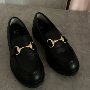 Svarta loafers med gulddetalj från XIT - Snygga svarta loafers från XIT med chunky platt sula och guldfärgad metallspänne över foten. Skorna har en rund tå och är tillverkade i slätt skinnliknande material. Perfekt för dig som gillar klassisk stil med en modern twist.