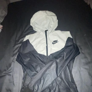 Nike svartvit vindjacka med huva - Nike vindjacka i svart och vitt med huva och hel dragkedja framtill. Jackan har ribbade muddar, två sidofickor med dragkedja och klassisk Nike-logga på bröstet. Perfekt för dig som gillar sportig och clean stil.