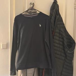 Mörkblå tröja från U.S. Polo Assn. - Snygg mörkblå tröja från U.S. Polo Assn. med klassisk rund hals och broderad logga på bröstet. Tröjan har långa ärmar och en mjuk känsla, perfekt för chill dagar. Diskret röd och vit rand på insidan av halsen ger en extra detalj.