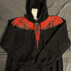 Svart hoodie Marcelo Burlon med röd print - Svart hoodie från Marcelo Burlon County of Milan med ikoniskt rött vingmönster över axlar och bröst. Tröjan har huva med snörning och långa ärmar. Materialet är mjuk bomull och passformen är normal. Snygg streetwear-vibe och riktigt fet design.