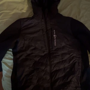 Svart pufferjacka från Sail Racing - Svart pufferjacka med huva från Sail Racing. Jackan har quiltad framsida, dragkedja och logga på bröstet. Materialet är syntet och polyester, perfekt för kyliga dagar. Långärmad och med en clean, sportig look.