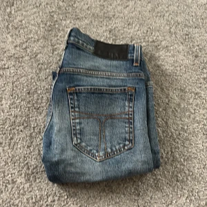 Blå jeans från Tiger of Sweden - Snygga blå jeans från Tiger of Sweden i klassisk denim. Modellen har fem fickor, orangea sömmar och en stilren svart patch bak i midjan. Passar dig som gillar en tidlös look och vill ha jeans med normal passform och raka ben.
