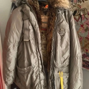 Parajumpers grå med päls - Parkajacka från Parajumpers i grått med fluffig päls runt huvan och foder i pälsimitation. Jackan har flera fickor, gul detalj vid dragkedjan och broderad patch på ärmen. Slim fit och perfekt för kalla vinterdagar. Är L men passar som M i storleken 