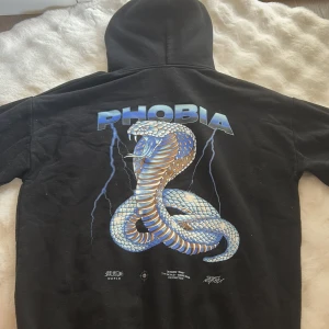 Svart hoodie Phobia med kobramotiv - Svart hoodie från Phobia med stor, detaljerad kobramålning och blixtar på ryggen. Framsidan har texten 'THE WORLD gone MAD' i ljusblått. Tröjan har huva och är långärmad, perfekt för dig som gillar streetwear med attityd.